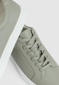 Adidași din piele texturată gri deschis, cu șireturiplate, tălpi albe și un accent dintr-un material neted pe lateral. Design clasic, de tip low-profile.