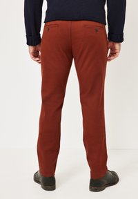 Rust-colored chinos with a straight-leg design, two back pockets with buttons, and a smooth, cotton material. Black shoes visible at the bottom.