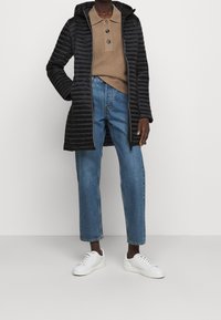 Schwarze Steppjacke mit einem Reißverschluss vorne und Kapuze. Getragen über einem sandfarbenen Polo und kombiniert mit hellblauen geraden Jeans und weißen Sneakern.