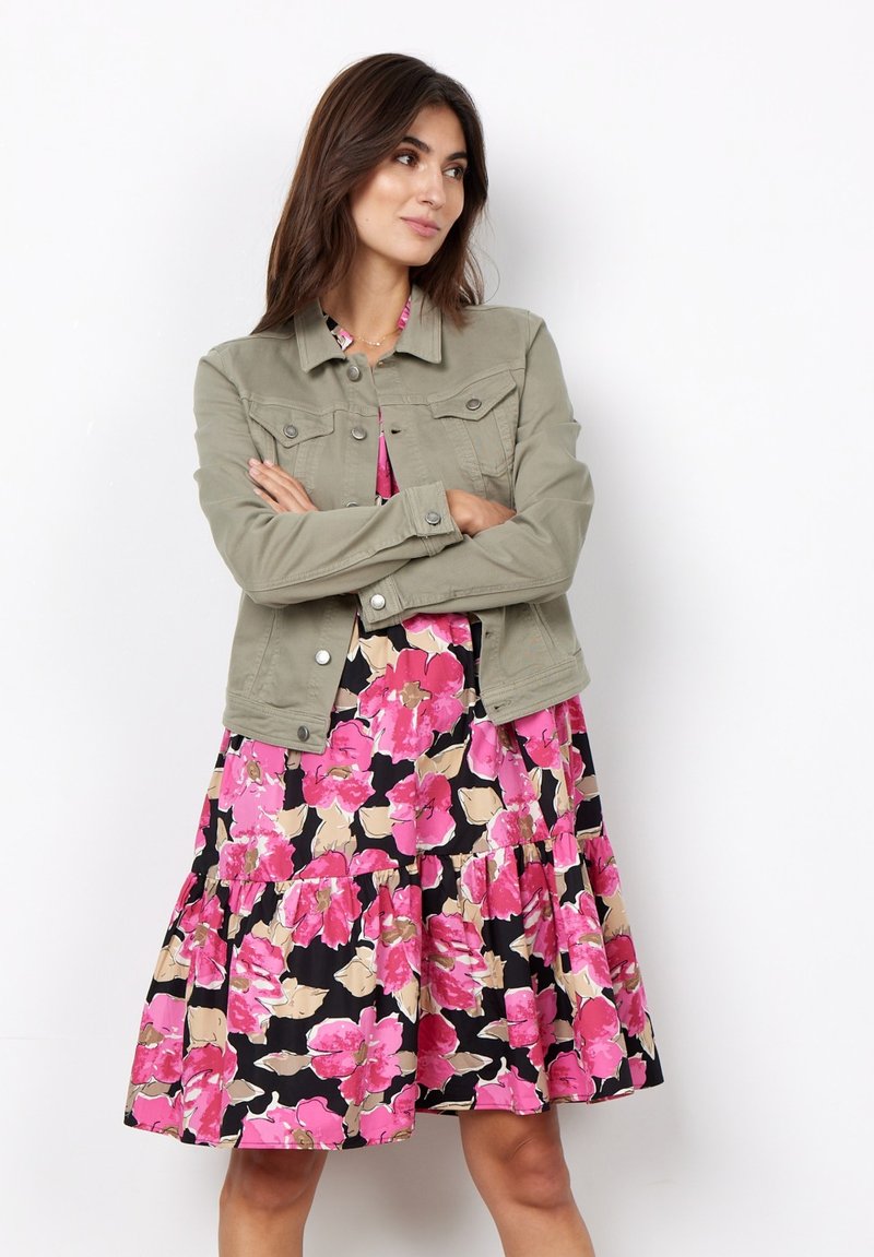 Lichtgroene denim jas met voorzakken, gelaagd over een zwarte jurk met grote roze en beige bloemenpatronen en een rimpelige onderrand.