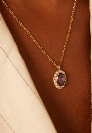 Collier en or avec un pendentif en pierre précieuse ovale violet entouré de strass transparents, sur fond de peau texturée.