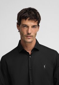 Zwarte button-up shirt gemaakt van gladde stof, met een subtiel geborduurd logo op de linkerborst en een klassiek kraagontwerp.