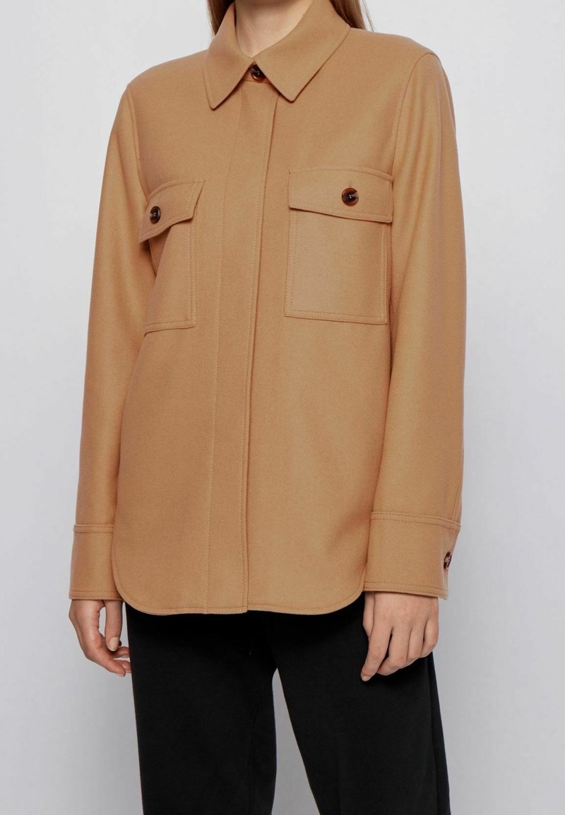 Personne portant une chemise camel à boutons avec deux poches poitrine et un pantalon noir, montrée des épaules aux hanches sur un fond uni.