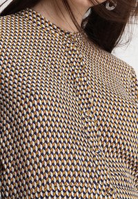 Musterbluse in Marineblau, Weiß und Senfgelb. Hat ein geometrisches Design, Knopfverschlüsse und einen runden Ausschnitt. Angenehme Stofftextur.