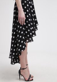 Femme portant une robe noire asymétrique à pois et des sandales noires à talons hauts, debout sur un fond clair.