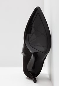 Chaussure à talon haut en daim noir avec un bout pointu et une semelle noire brillante. Elle présente un accent zippé et un design élégant et épuré.