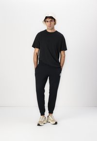 Tricou negru din bumbac, pantaloni scurți negri cu logo, pălărie tip bucket crem și adidași bej. Design simplu, croi lejer și stil minimalist.