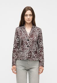 Γυναίκα που φοράει ένα εφαρμοστό σακάκι με λεοπάρ μοτίβο και δύο κουμπιά, συνδυασμένο με ανοιχτό γκρι παντελόνι, στέκεται μπροστά σε λευκό φόντο.