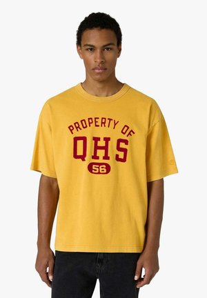 Giovane uomo che indossa una maglietta gialla con la scritta rossa "PROPERTY OF QHS 56", in piedi davanti a uno sfondo bianco uniforme.