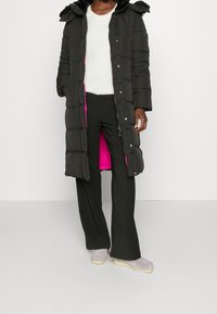 Černý prošívaný dlouhý kabát s umělou kožešinou na kapuci, jasně růžovou podšívkou a zapínáním na patenty/tlačítka. V kombinaci s černými kalhotami a světle šedými botami.