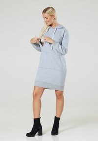 Grauäugiges Kleid aus weichem Material mit einer Fronttasche und kontrastierendem Saum. Kombiniert mit schwarzen, mit Absatz versehenen Stiefeletten. Glatte Textur.