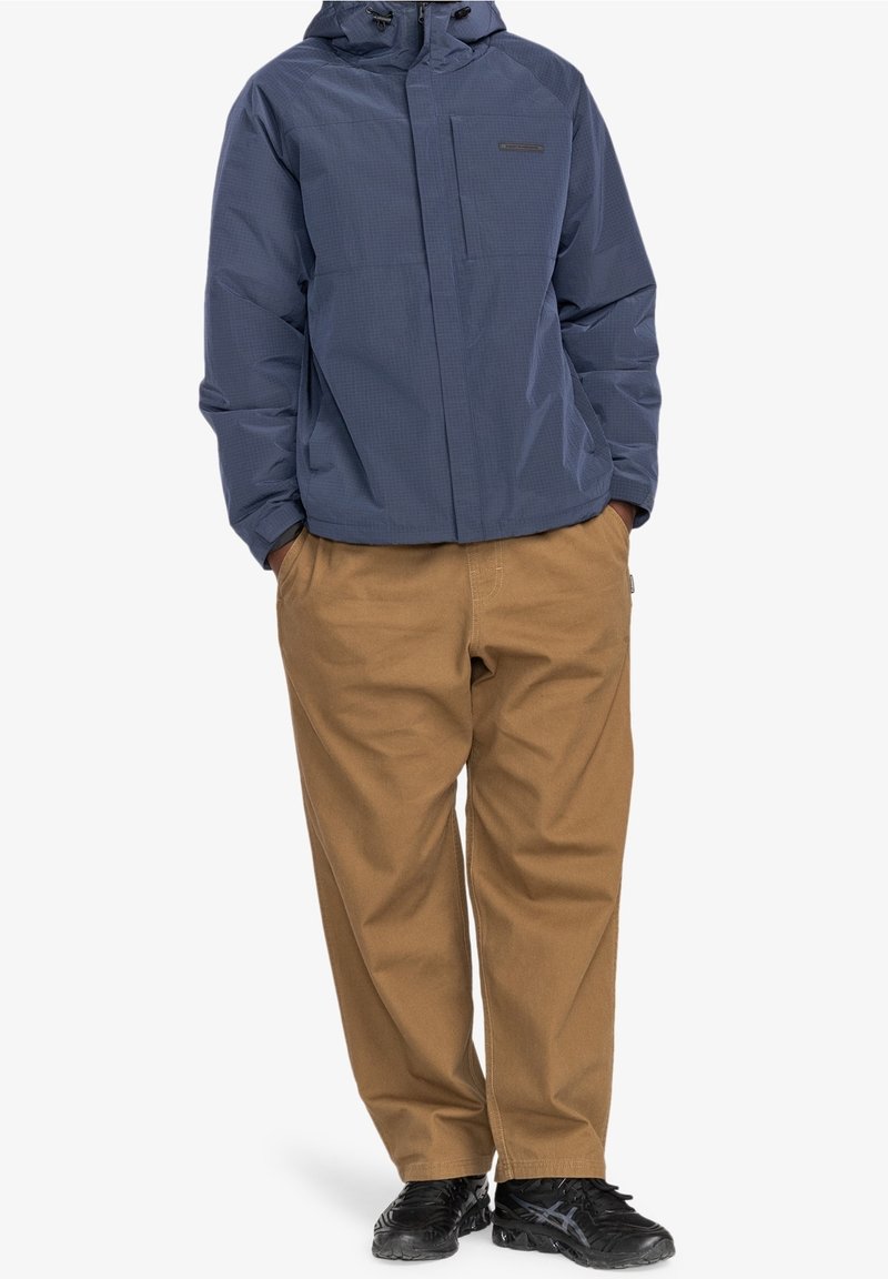 Granatowa ocieplana kurtka z kapturem, o fakturowanej powierzchni i przedniej kieszeni. Połączona z beżowymi spodniami o szerokich nogawkach i czarnymi butami.