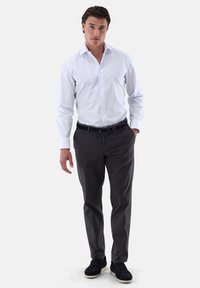 Camicia bianca a righe con maniche lunghe e colletto, abbinata a pantaloni grigio scuro e scarpe blu navy. Il modello sta in piedi con le mani in tasca.
