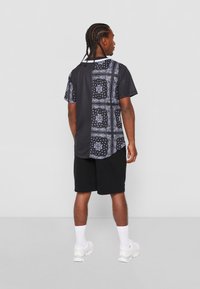 Kortærmet sort skjorte med et bandana-mønster i blå nuancer på ryggen, parret med sorte shorts og hvide sneakers.