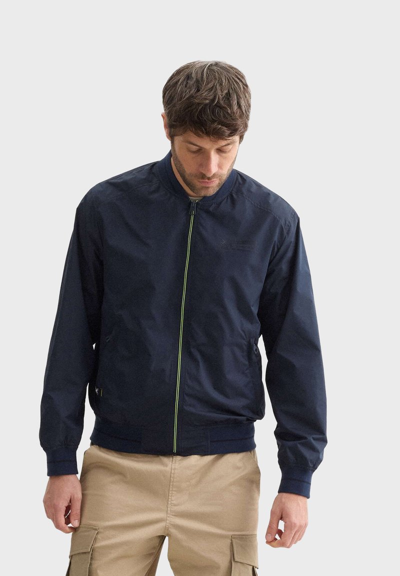 Námořnická bomber bunda s předním zipem, dlouhými rukávy a bočními kapsami. Obsahuje živou zelenou akcentu podél zipu. Lehká tkanina.