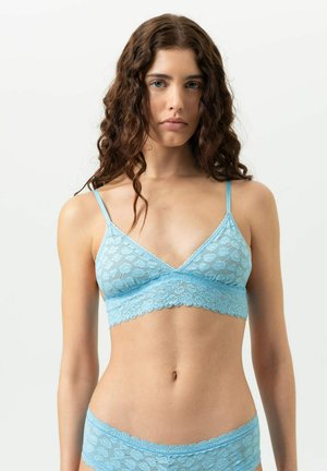 Jonge vrouw met golvend bruin haar, gekleed in een lichtblauwe kanten bralette en bijpassend ondergoed, staand tegen een effen witte achtergrond.