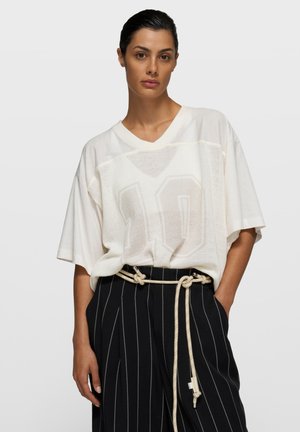 Kobieta z zaczesanymi do tyłu włosami, ubrana w luźną białą siateczkową bluzkę z nadrukiem "10" oraz czarne spodnie w prążki z sznurkiem zawiązanym w supeł jako paskiem, z rękami w kieszeniach.