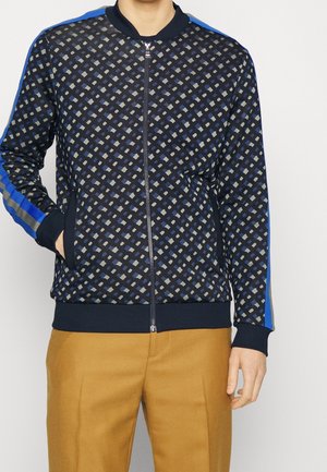 Homme portant une veste bleu marine zippée avec un motif en losanges gris et bleu clair et des rayures bleues aux manches, associée à un pantalon moutarde.