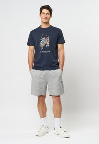 Tamsiai mėlynas medvilnės marškinėliai su polo grafika, derinami su pilkais džersiniais šortais. Šortai turi šonines kišenes ir logotipo detalę. Balti sportiniai bateliai ir raitos kojinės.