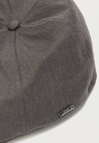 Γκρι καπέλο τύπου flat cap από υφασμάτινο ύφασμα με υφή ψαροκόκκαλο, με στρογγυλεμένο σχήμα, ραφές και μια μικρή μεταλλική ετικέτα με λογότυπο.