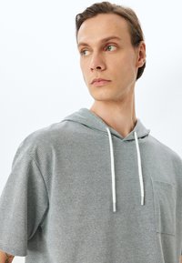 Sudadera gris con un bolsillo frontal, cortes suelto, mangas cortas y cordones blancos. La tela parece suave con un acabado texturizado.