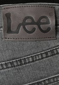 Tessuto denim grigio con un'etichetta in pelle con il logo "Lee" impresso in nero. Cuciture visibili e dettagli della trama sul tessuto.