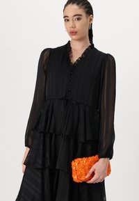 Black layered dress with sheer sleeves and ruffled neckline, featuring a tie waist. Holding a textured orange sequin clutch purse.