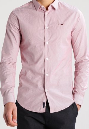 Mand iført en tætsiddende lyserød skjorte med knapper og krave samt en lille "AJ"-logo på venstre bryst, kombineret med mørke bukser.