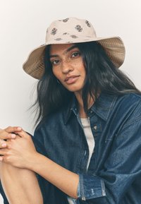 Beige Blumen-Eimerhut mit breiter Krempe, aus leichtem Stoff gefertigt. Verfügt über ein filigranes Blumenmuster in dunklen Farben.