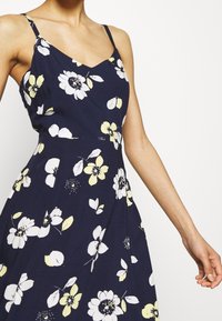 Marineblaues Kleid mit weißen und gelben Blumenmuster, Spaghetti-Trägern, V-Ausschnitt und einem fließenden, knielangen Design. Sanfte Stofftextur.