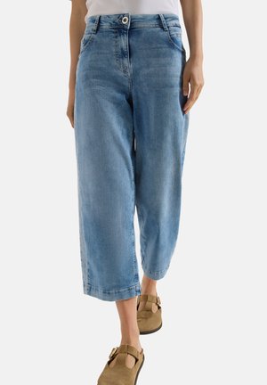 Personne portant un jean large court bleu clair et des sandales marron en daim avec boucle, avec un haut blanc visible à la taille.
