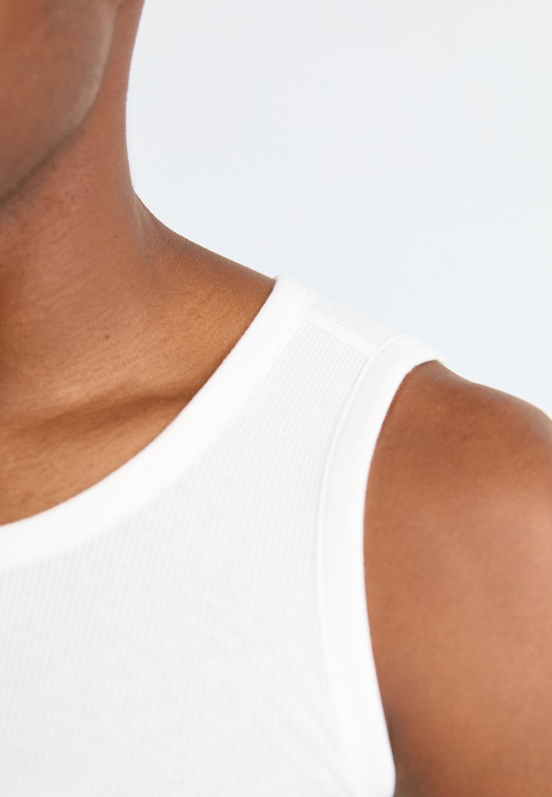 Nahaufnahme des Kragens eines weißen gerippten Tanktops, das den Ausschnitt, die glatte Textur und das weiche Baumwollmaterial hervorhebt. Teilweise sichtbarer Hautton.