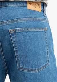 Blauwe denim jeans hebben oranje stiksels, een leren label op de achterste tailleband en een enkele achterzak aan de rechterzijde.