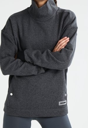 Person i en mørkegrå rullekravetrøje med armene over kors, iført grå leggings, står mod en ensartet lys baggrund.