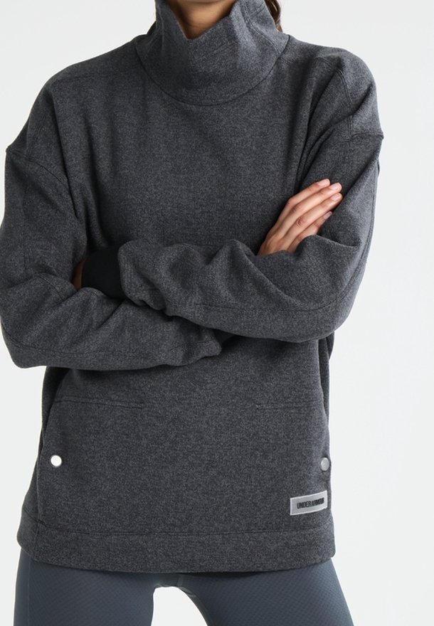 Personne en sweat à col roulé gris foncé, les bras croisés, portant des leggings gris, debout devant un fond clair et uni.