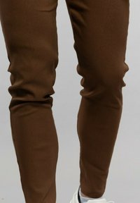 Pantalon slim marron en tissu lisse, avec un design épuré et une coupe fuselée, associé à des baskets blanches.