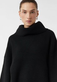 Czarny sweter z prążkowanego dzianiny z wysokim, szerokim kołnierzem i luźnym krojem, wykonany z teksturowanej tkaniny i z długimi rękawami. Prosty design bez wzorów.