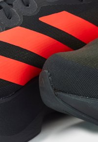 Chaussures de sport noires avec rayures rouges, dessus en tissu texturé et bout arrondi. Comprend une semelle en caoutchouc et un logo de marque distinct sur le talon.