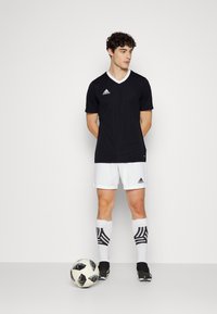 Svart fotbollströja med vit V-hals, tillsammans med vita shorts och randiga svarta strumpor som går upp över vaden, stående bredvid en fotboll.