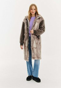 Žena v dlouhém hnědém tečkovaném umělém kožichu, pod ním fialový svetr, modré džíny a černé platformové boty, stojící na bílém pozadí.