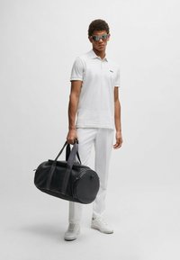 Bela polo majica s teksturiranim dizajnom, crni logotip; elegantna crna torba na točkiće sa sivim trakama; model u belim pantalonama i belim patikama.