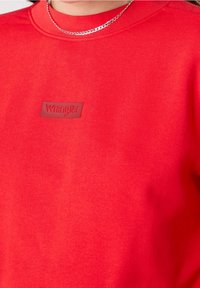 Sweatshirt rouge en tissu texturé, avec un patch logo en caoutchouc "Wrangler" au centre. Encolure arrondie et détails de couture visibles.