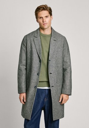 Cappotto grigio a tweed con colletto a punte, design a un petto e bottoni blu, indossato sopra un maglione verde e jeans di denim.