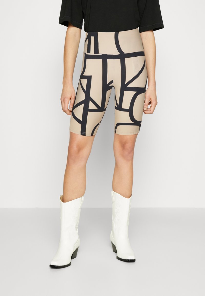 Personne portant un short de cyclisme beige avec des lignes abstraites noires, un haut noir et des bottes de cowboy blanches, debout sur un fond blanc.