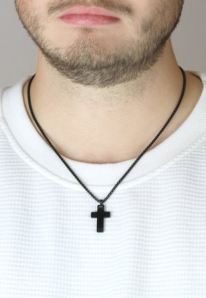 Schwarzes Kreuz-Anhänger an einer schwarzen Kette, getragen über einem strukturierten weißen Pullover. Das Kreuz hat eine glatte Oberfläche und ist zentriert positioniert.
