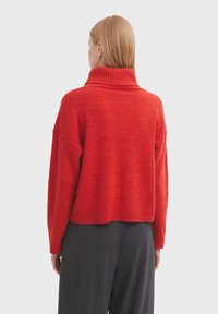 Roter, strukturierten Rollkragenpullover mit einem verkürzten Design und geripptem Kragen. Das Model trägt ihn mit schwarz gestreiften Hosen, von hinten gezeigt.