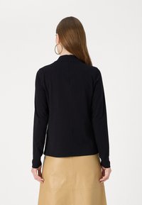 Svart langermet topp med høy hals, med en skjult glidelås bak og slank passform, kombinert med et beige skjørt.