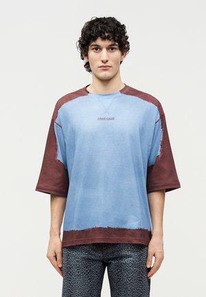 Νεαρός άνδρας με σγουρά μαλλιά, φορώντας μπλε και μπορντό πουκαμίσα με κοντά μανίκια σε υπερμεγέθη μέγεθος και σκούρο παντελόνι με σχέδια, στέκεται μπροστά σε απλό φωτεινό φόντο.