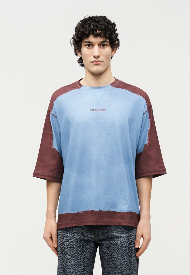 Νεαρός άνδρας με σγουρά μαλλιά, φορώντας μπλε και μπορντό πουκαμίσα με κοντά μανίκια σε υπερμεγέθη μέγεθος και σκούρο παντελόνι με σχέδια, στέκεται μπροστά σε απλό φωτεινό φόντο.