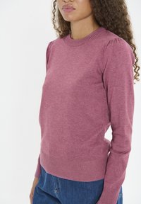Maglione rosa chiaro realizzato in tessuto morbido, con collo rotondo, polsini a costine e maniche leggermente a sbuffo. Abbinato a jeans blu.